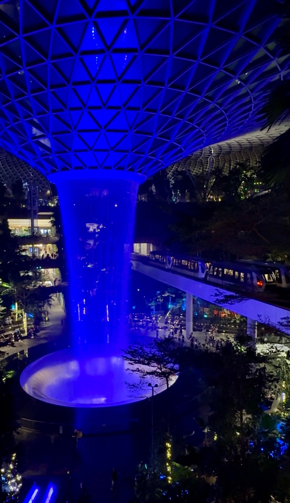 The Rain Vortex indoor waterfall glowing under the geometric glass dome of Jewel Changi Airport in Singapore at night, with lush greenery and an elevated monorail track above.