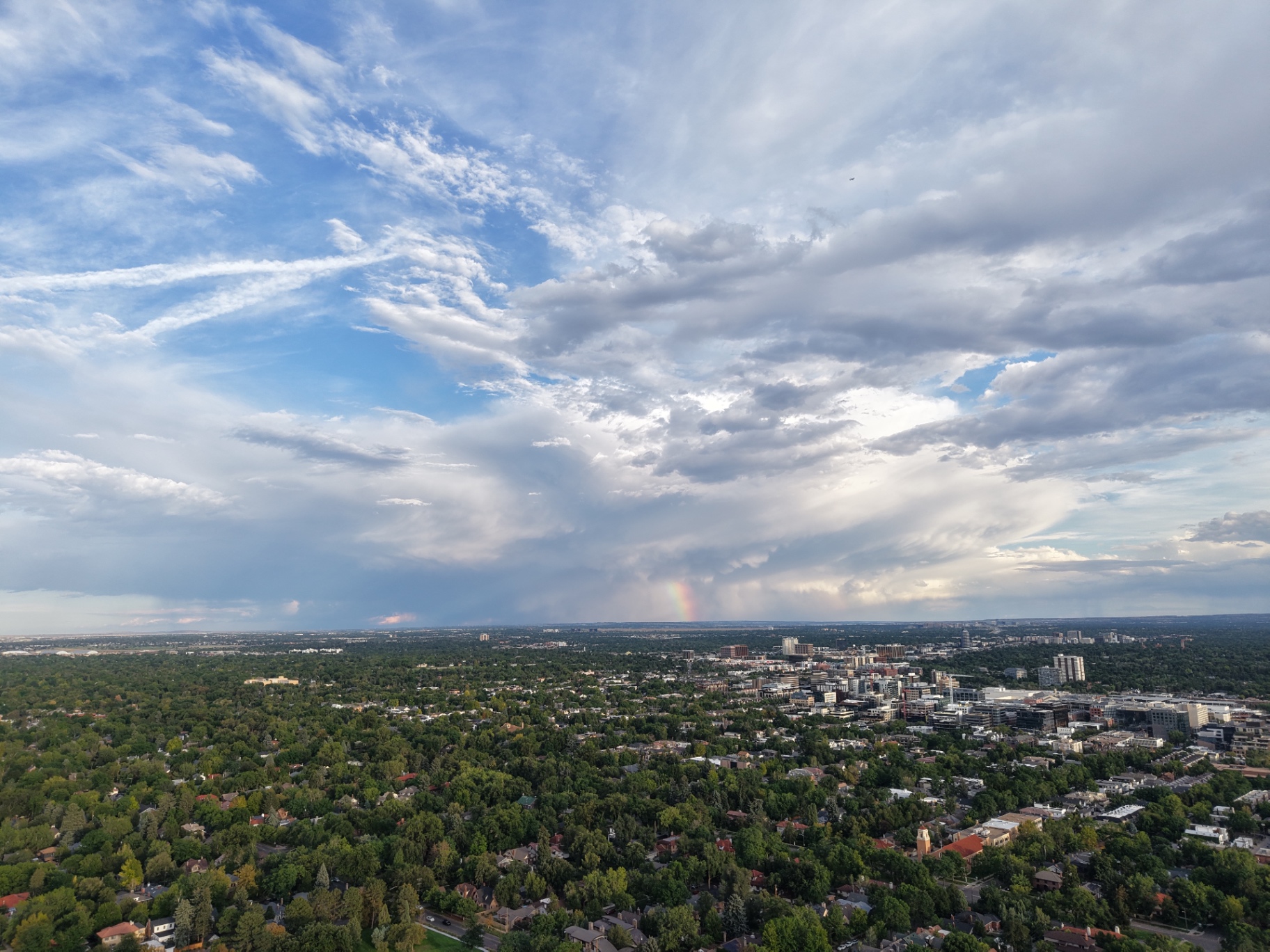 Aerial view over the tree-filled neighbourhoods and skyline of Denver, Colorado, with a rainbow beneath a dramatic bank of storm clouds.