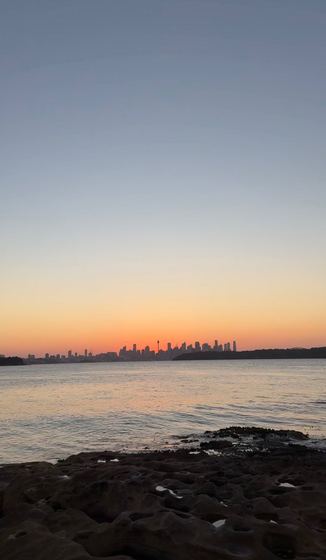 The Sydney skyline at dusk seen across the water from a rocky shoreline, with the Sydney Tower Eye rising above the high-rises and sunset light on the harbour.