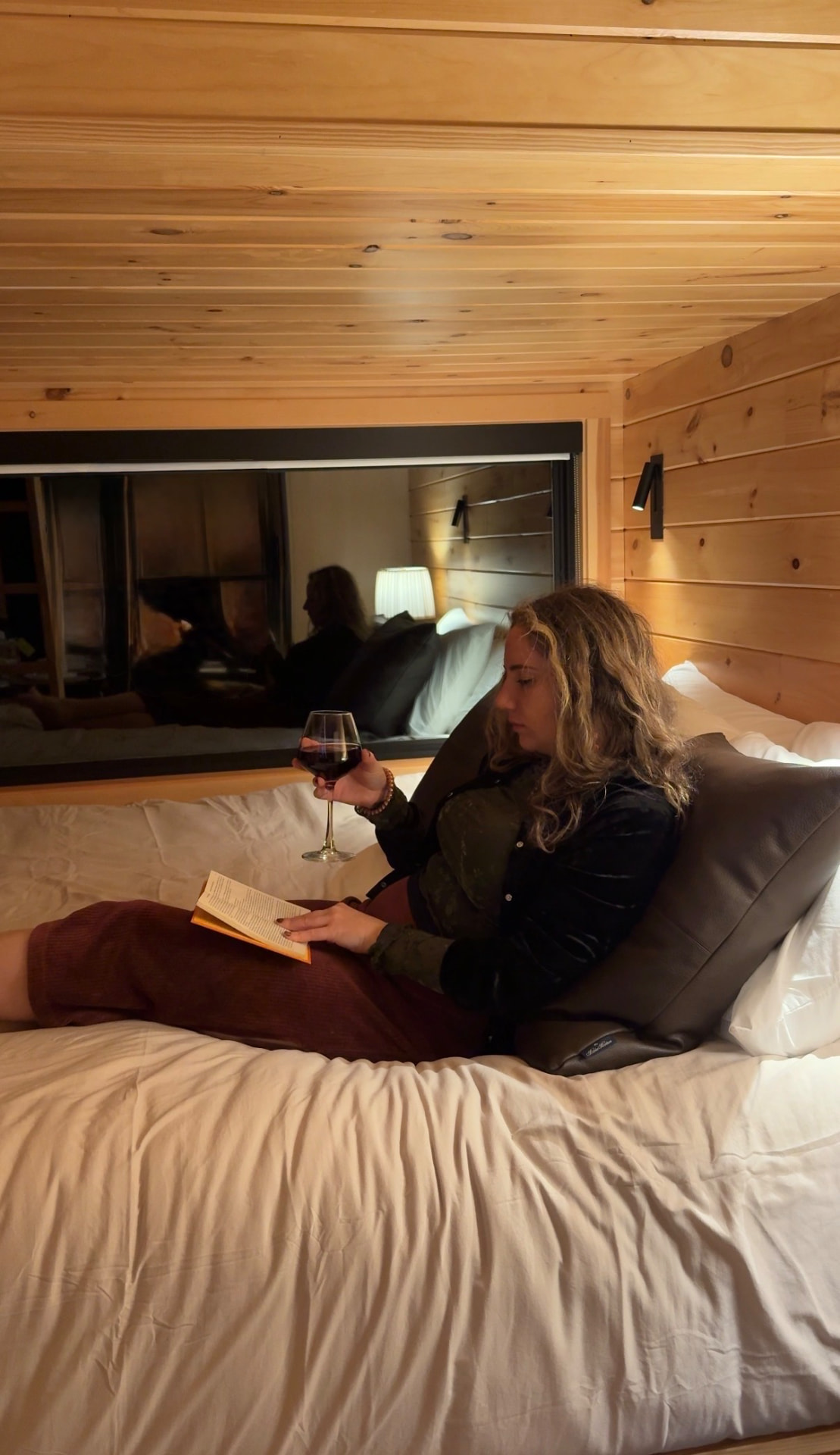 Miranda reading a book with a glass of red wine on the bed of a wood-paneled cabin at The Drop Inn Knoxville