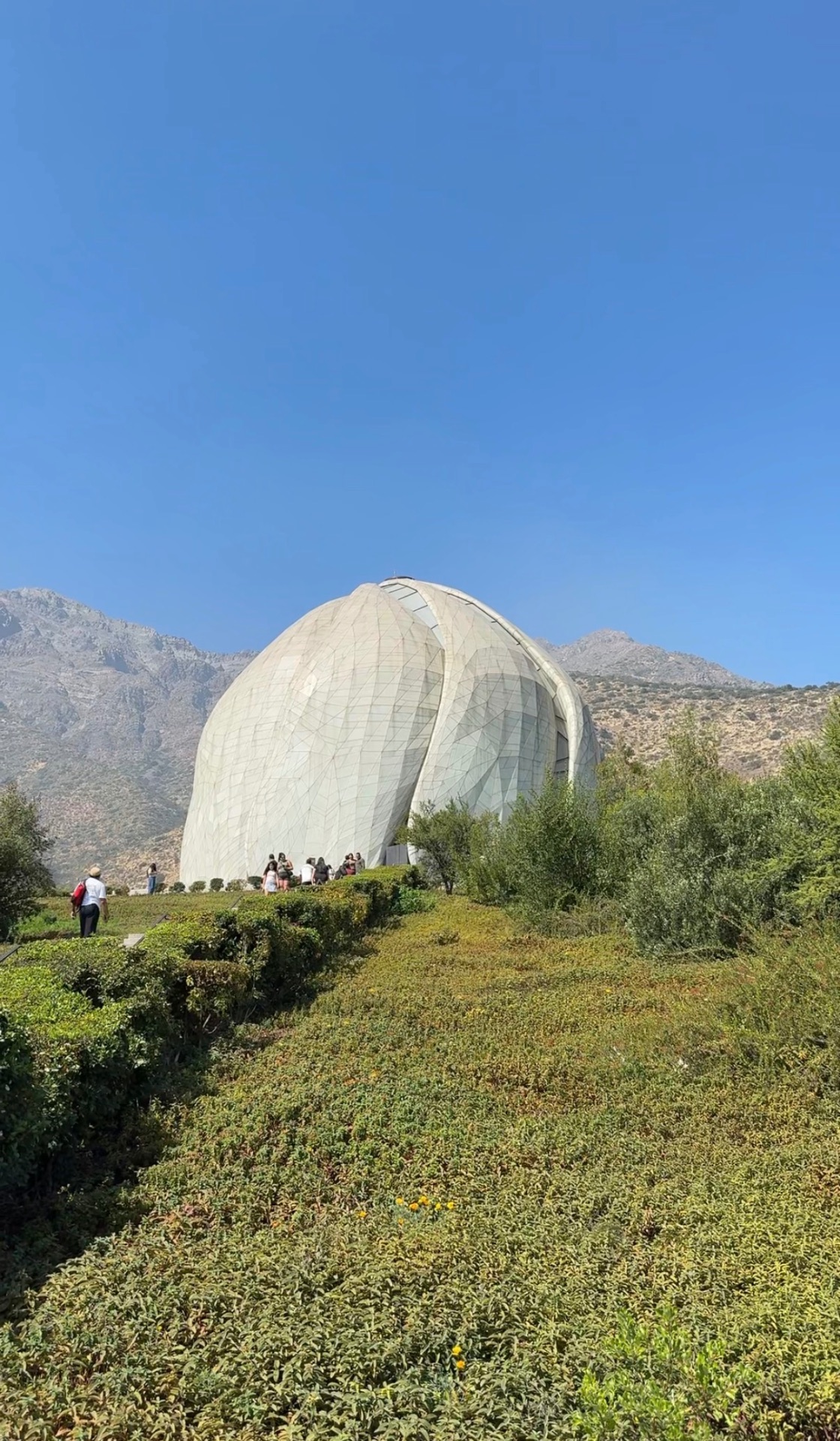 The Bahá'í Temple of South America in Santiago, its nine translucent petal-shaped panels rising above trimmed hedges and a paved path, with the arid Andean foothills behind under a clear blue sky.