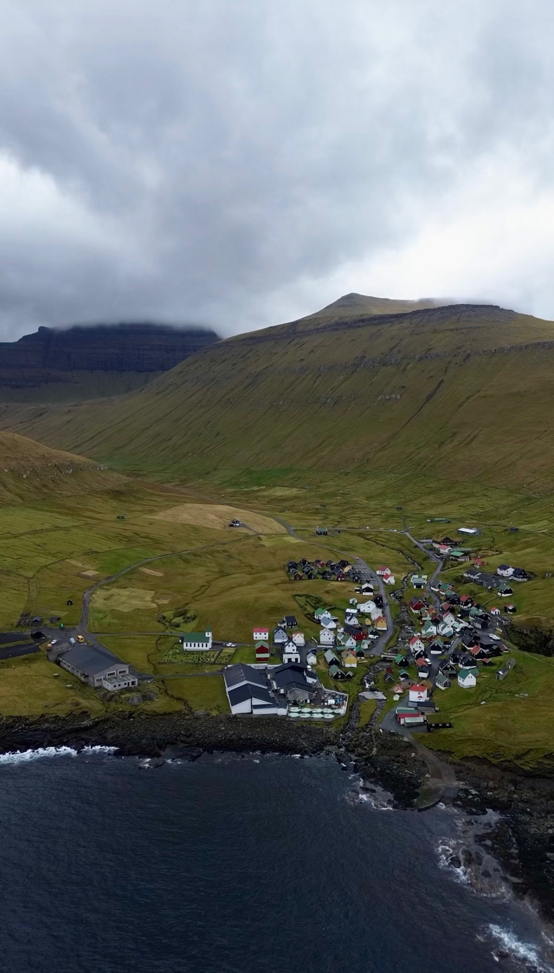 Aerial view of a small Faroese village of colourful houses beside the Atlantic, backed by steep green mountains under low cloud.