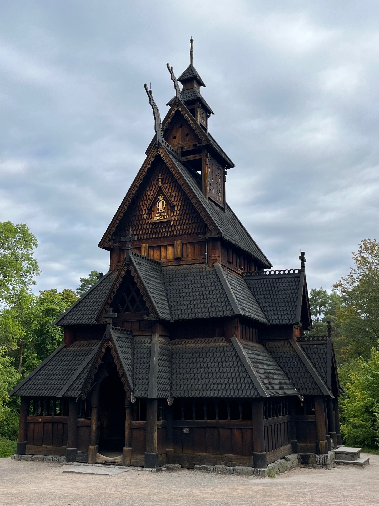 The dark wooden Gol Stave Church in Oslo, Norway, with its steep shingled roofs, carved dragon-head gables and bell tower rising against a cloudy sky, framed by green forest.