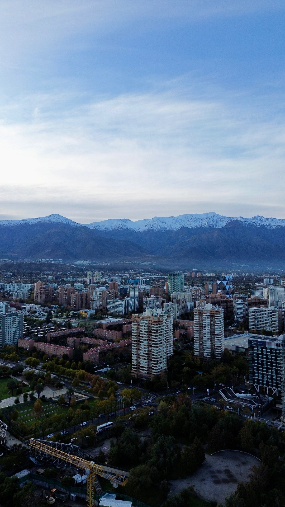 Aerial view of high-rise apartment towers and parkland in Santiago, Chile, at dusk, backed by the snow-covered Andes mountains.