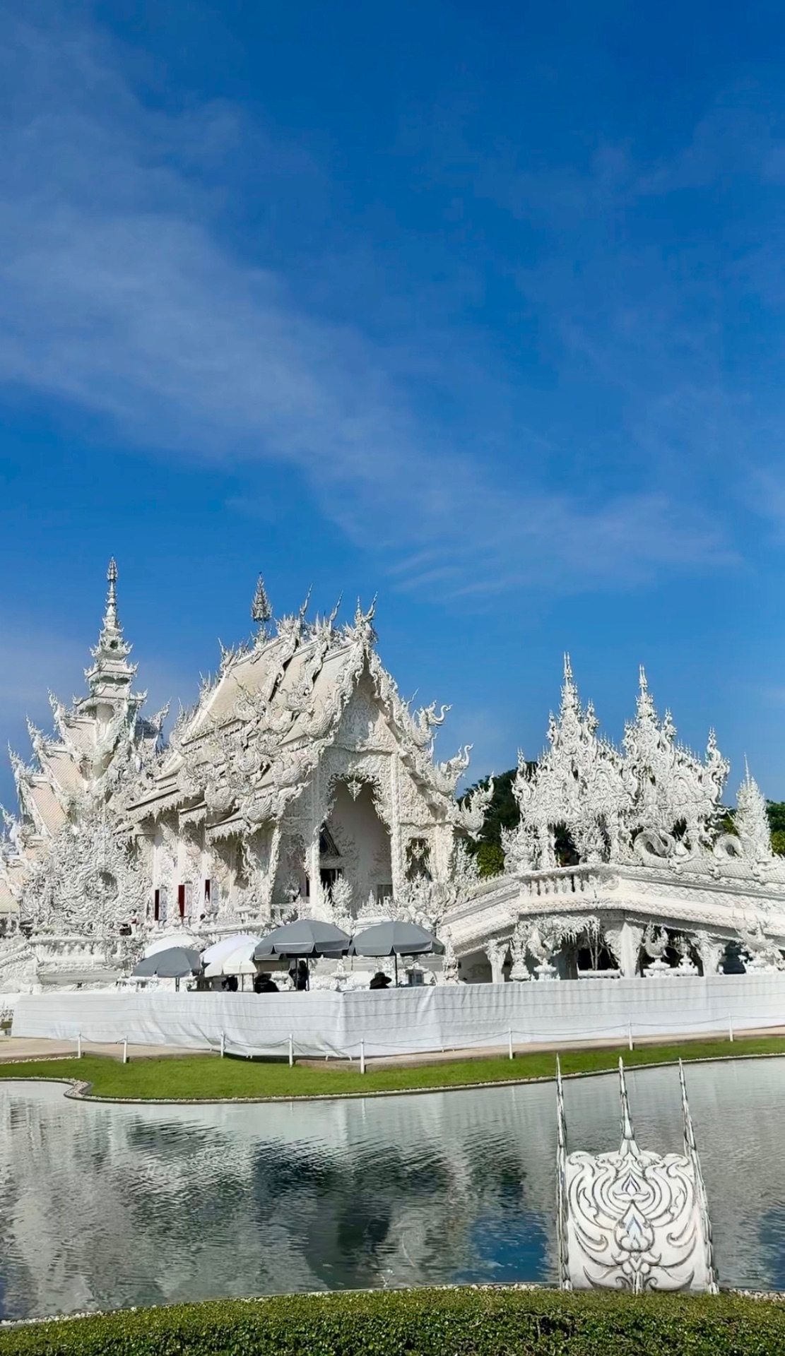 Wat Rong Khun, the White Temple in Chiang Rai, with ornate white carvings reflected in the pond under a blue sky