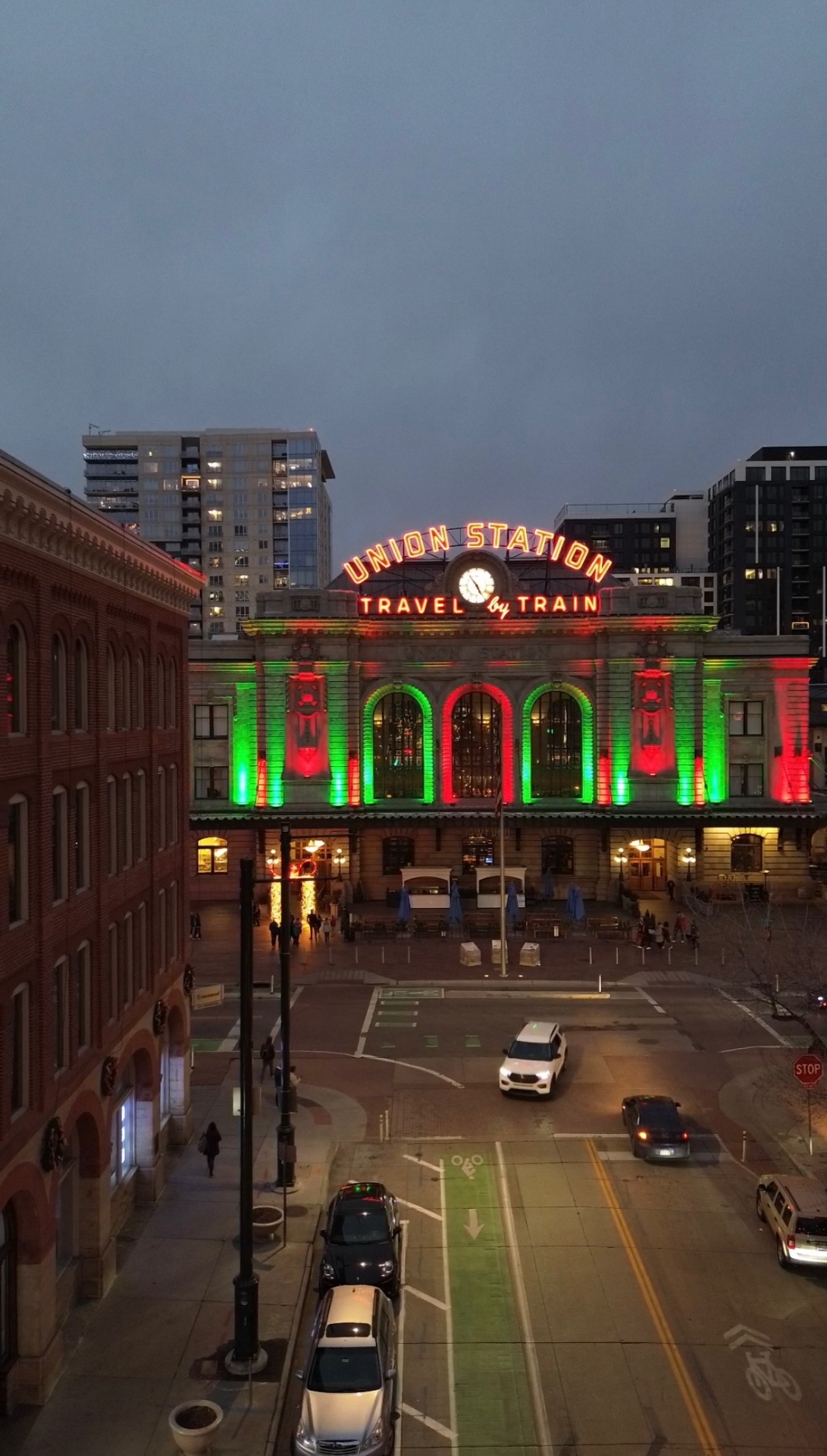 Denver Union Station lit in red and green at dusk, with its neon 'Travel by Train' sign glowing above the street.